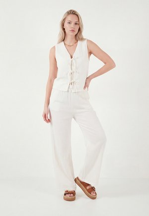 Blonde Frau trägt ein ärmelloses weißes Top mit vorderer Knotenbindung, lockere weiße Hose, braune Plateausandalen, steht mit einer Hand in der Hüfte.