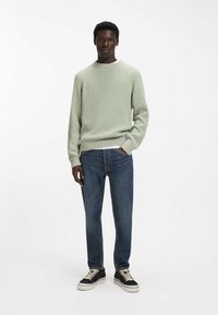 Jasnozielony prążkowany sweter z długimi rękawami, okrągłym dekoltem i fakturowanym wykończeniem, zestawiony z ciemnoniebieskimi jeansami o prostych nogawkach i czarnymi sneakersami.