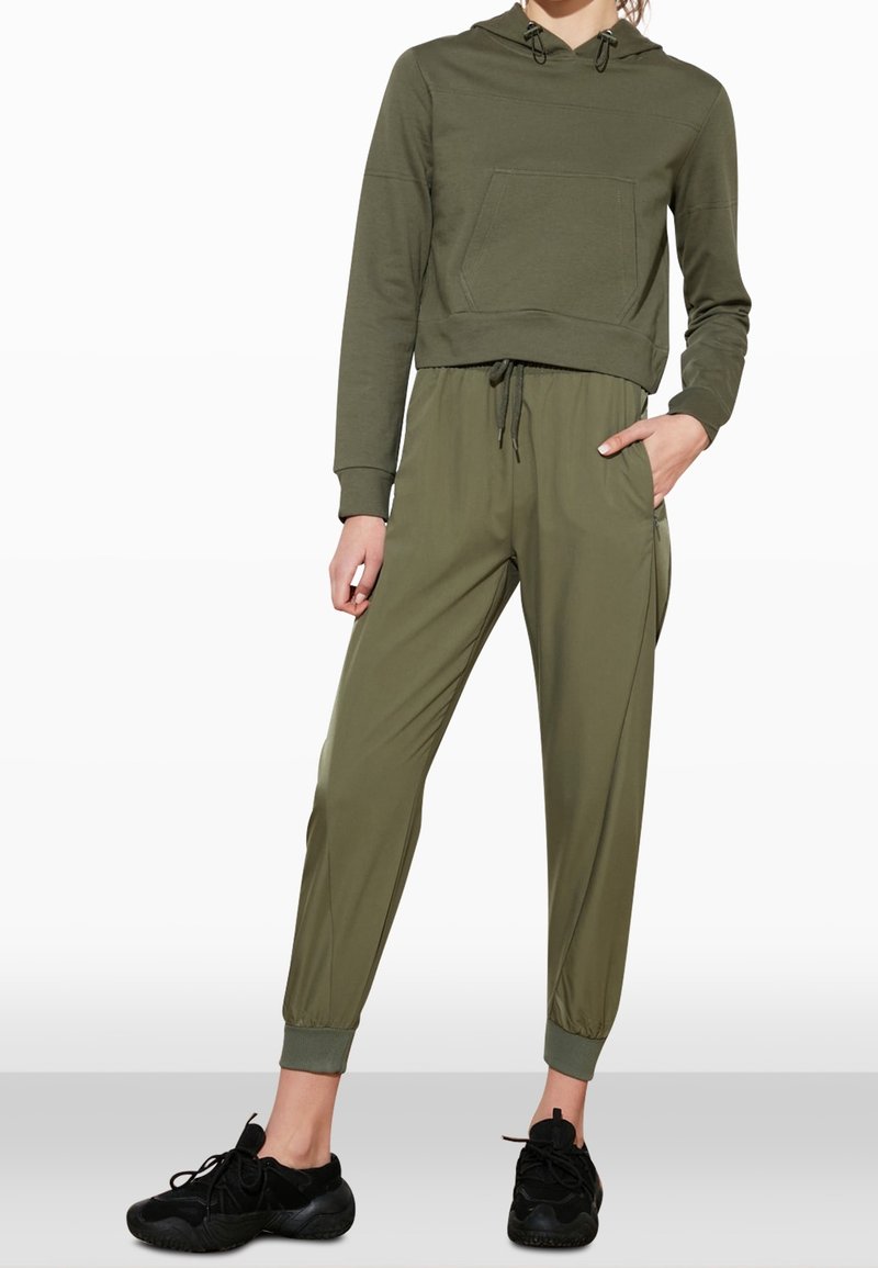 Personne portant un sweat à capuche court vert olive, un pantalon de jogging assorti avec cordon de serrage et chevilles resserrées, et des chaussures de sport noires, debout avec une main dans la poche.