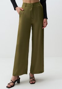 Oliwkowe spodnie z szerokimi nogawkami z gładkiego materiału z bocznymi kieszeniami. Posiadają wysoki stan i subtelne fałdy; zestawione z czarnymi sandałami na obcasie.