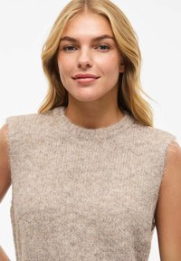 Blondīne sieviete, kas valkā bezroku bēša krāsas adītu džemperi, viegli smaida, ar vaļīgiem matiem uz vienkrāsaina balta fona.