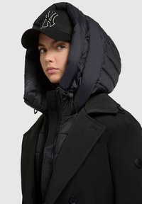 Vêtements d'extérieur matelassés noirs avec une grande capuche structurée et une casquette noire portant un logo blanc. La veste a un devant à double boutonnage et des zips.