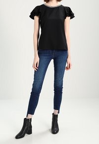 Black short-sleeve top with ruffle accents and a decorative neckline paired with skinny blue jeans and black ankle boots.