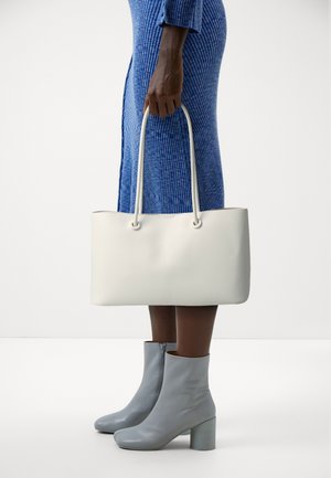 Kobieta w niebieskiej, prążkowanej sukience trzyma prosta, białą, prostokątną torebkę, nosząc jasnoszare botki do kostek na słupku z bocznymi zamkami.