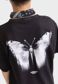 Schwarzes T-Shirt mit einem großen weißen Schmetterlingsdesign auf dem Rücken, mit einer strukturierten Oberfläche und dem Text "REDEFINED REBEL" am unteren Rand.