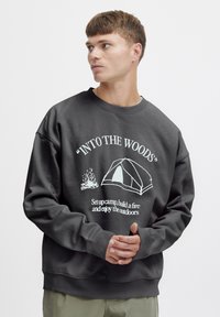 Sudadera de un gris oscuro con cuello Redondo, que presenta texto gráfico blanco e ilustraciones de una tienda de campaña y una hoguera. Ajuste suave y relajado.