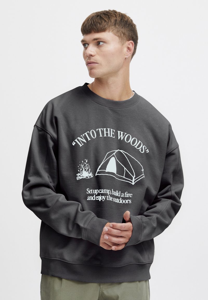 Sudadera de un gris oscuro con cuello Redondo, que presenta texto gráfico blanco e ilustraciones de una tienda de campaña y una hoguera. Ajuste suave y relajado.