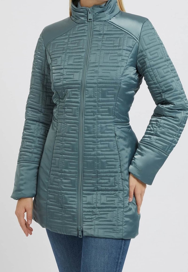 Chaqueta acolchada de color azul verdoso con un cuello alto, que presenta un patrón geométrico, un cierre frontal con cremallera y bolsillos laterales. Tejido suave con un ligero brillo.