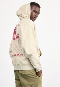Sudadera beige con un gran gráfico burdeos en la parte posterior, que presenta texto y coordenadas. Tejido de mezcla de algodón con una textura suave y capucha.