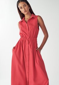Czerwona sukienka bez rękawów z kołnierzykiem, z przodu zapinana na guziki, z elastycznym pasem i kieszeniami. Wykonana z lekkiego materiału, o zwiewnym kroju.
