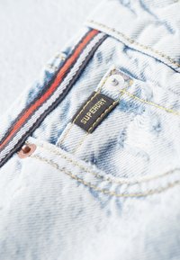 Poche en denim bleu clair avec un design à rayures noires et rouges, des coutures jaunes, une étiquette de marque et un détail de rivet en cuivre.
