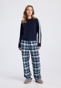 Navy knit sweater with white stripes on sleeves, paired with blue and white plaid pajama pants, and beige slippers. Soft textures visible.