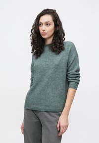 Svetlo zelena pulover s rebrastim vzorcem, okroglim izrezom in dolgimi rokavi, kombiniran s sivimi hlačami, ki izstopajo s svojo mehko teksturo in sproščenim krojem.