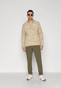 Maglione in pile beige con mezza zip e motivo astratto, abbinato a pantaloni verde oliva e sneakers bianche. Il modello indossa occhiali da sole.