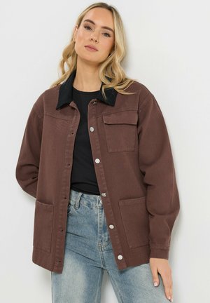 Kobieta ubrana w brązową, zapinaną na guziki kurtkę z czarnym kołnierzem, czarną koszulę i jasnoniebieskie dżinsy, stojąca na białym tle.