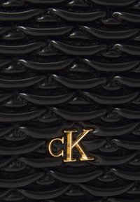 Matière texturée noire présentant des motifs ondulés et un logo "cK" doré proéminent au centre.