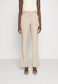 Femme portant un pantalon beige à taille haute à rayures avec des poches latérales, un haut en tricot sans manches blanc et des escarpins à bout pointu en peau de serpent.