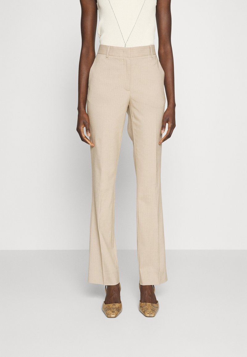 Femme portant un pantalon beige à taille haute à rayures avec des poches latérales, un haut en tricot sans manches blanc et des escarpins à bout pointu en peau de serpent.