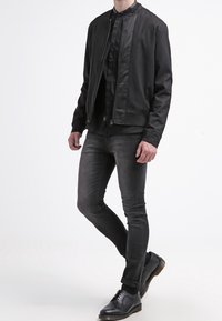 Giubbotto bomber nero con una finitura liscia, abbinato a jeans scuri aderenti e scarpe nere stringate. Design semplice e elegante; nessun logo visibile.