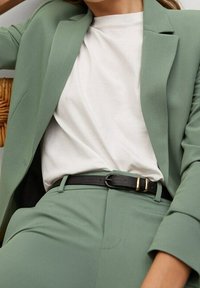 Zielony, dopasowany blezer noszony na białej koszuli, w zestawie z dopasowanymi zielonymi spodniami. Czarny pasek z złotymi akcentami podkreśla talię.