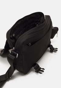 Borsa a tracolla nera in tessuto durevole, con più scomparti con cerniera, tracolla regolabile e dettagli in metallo a forma di artiglio.