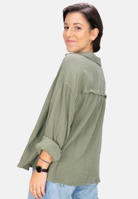 Olive green, textured button-up shirt with a collar, relaxed fit, and rolled sleeves. Worn with light blue jeans and a black watch.
