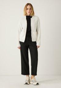 Weißer flauschiger Cardigan mit offenem Vorderteil, zwei frontalen Taschen, kombiniert mit einem schwarzen Oberteil und schwarzen weitgeschnittenen Hosen. Das Modell trägt cremefarbene und schwarze Sneakers.