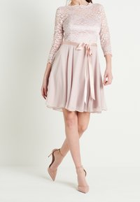 Woman wearing a knee-length pale pink dress with lace top, satin waist ribbon, and chiffon skirt, paired with nude ankle-strap high heels.