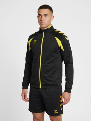 Ung mand iført en sort og gul hummel sportjakke og matchende shorts, der står mod en ensfarvet lys baggrund.