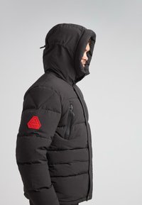 Mand iført en sort polstret vinterjakke med hætte oppe og et rødt logo-patch på venstre ærme mod en neutral baggrund.