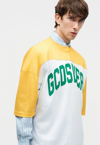 Νέος άνδρας που φοράει μια λευκή και κίτρινη υπερμεγέθη φανέλα GCDS πάνω από ένα μπλε ριγέ πουκάμισο, κοιτάζοντας ελαφρώς προς το πλάι.