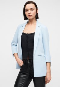 Νεαρή γυναίκα με ίσια σκούρα μαλλιά φοράει ανοιχτό μπλε σακάκι πάνω από μαύρο μπλουζάκι με κουμπιά και μαύρο δερμάτινο παντελόνι, στέκεται μπροστά σε απλό φόντο.