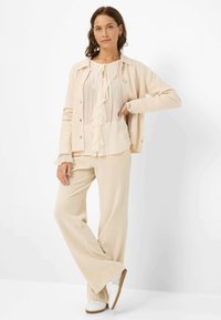 Beige ribbed cardigan worn over a light cream ruffled blouse, paired with wide-leg beige pants and white sneakers, set against a plain background.