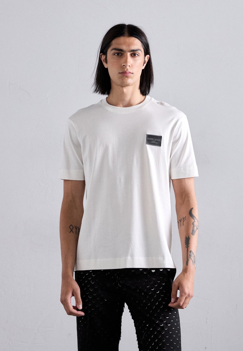 White cotton t-shirt with a black rectangular patch on the left chest. Short sleeves, round neckline, and a relaxed fit. Black patterned pants.