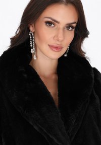 Mujer con cabello castaño que lleva grandes pendientes colgantes de perlas y cristales, vestida con un abrigo de piel sintética negro, con una ligera sonrisa.