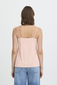 Ženska s rjavimi lasmi dolžine do ramen, oblečena v svetlo rožnato zgornji del na tanke naramnice in svetlo modre kavbojke, stoji obrnjena stran.