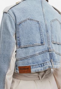 Kurtka dżinsowa z jasnoniebieskiego materiału, krótki fason, brązowa skórzana metka z napisem "Desigual" i premium denim, kontrastowe beżowe rękawy.
