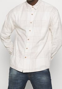Langarm katoenen overhemd in lichtbeige met subtiel ruitpatroon, met een button-down kraag en één borstzak. Gestyled met denim.