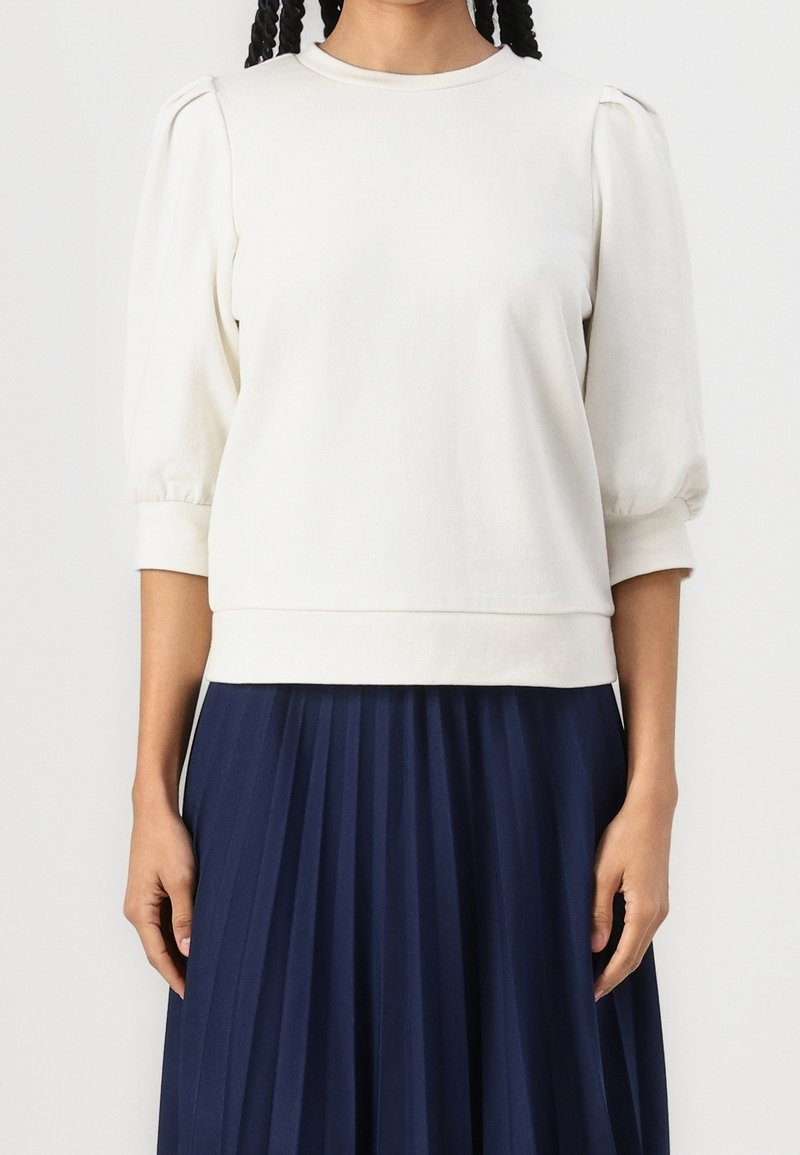 Femme portant un haut blanc à manches bouffantes trois-quarts associé à une jupe plissée bleu marine, debout devant un fond uni.