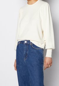 Felpa color crema con maniche arricciate, scollo rotondo e un motivo strutturato sulla parte superiore. Abbinata a jeans in denim blu a vita alta.