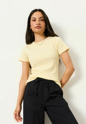 Femeie cu păr lung și închis la culoare, purtând un tricou galben pal cu mânecă scurtă și pantaloni negri cu șnur, stând în fața unui fundal alb.
