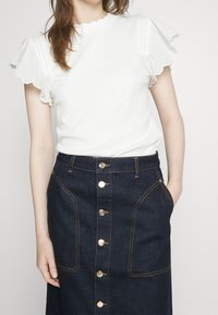 Vit kortärmad topp med scallopdetaljer, kombinerad med en mörk denimkjol med guldknappar och framfickor.