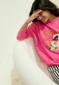 Sudadera rosa con gráfico de "Fresh Up" y diseño de 7-Up, con mangas largas y puños acanalados. Usada con pantalones a rayas, sentado.