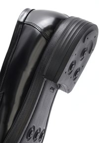 Zwarte formele leren schoen met gestikte details en een gestructureerde rubberen zool met cirkelvormige grip patronen en kleine gaatjes.