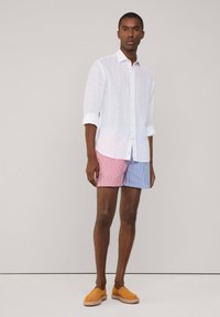 Camicia di lino bianca con maniche arrotolate, abbinata a pantaloni corti a righe rosse e blu. Modello che indossa scarpe slip-on beige, in piedi davanti a uno sfondo neutro.