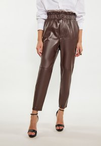 Bruine neplederen broek met een elastische tailleband, taps toelopende benen en een glanzende afwerking, gecombineerd met zwarte bandjeshakken.