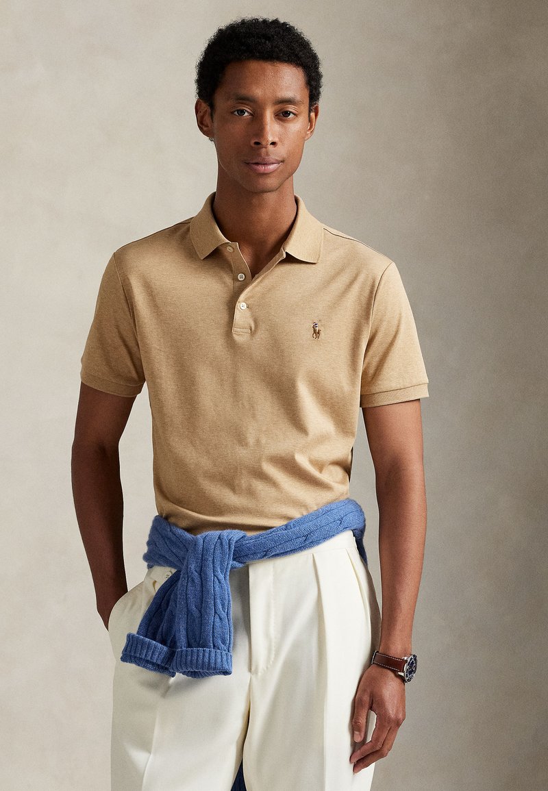 Tânăr purtând un tricou polo bej cu mânecă scurtă, pantaloni albi, un pulover albastru legat în jurul taliei și un ceas de mână, stând cu mâna în buzunar.