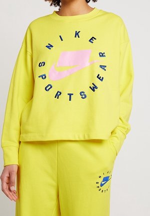Persona che indossa un vivace completo sportivo Nike giallo con un logo circolare rosa e azzurro sulla felpa e sui pantaloni, con una mano in tasca.