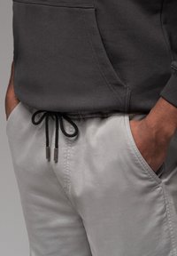 Grijze katoenen hoodie met een voorzak, gecombineerd met lichtgrijze broek met koordjes en metalen tips. De textuur is zacht.