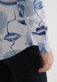 Chemise blanche avec des motifs de visage abstraits en bleu, dotée d'un poignet boutonné et d'une texture lisse, associée à un pantalon sombre.